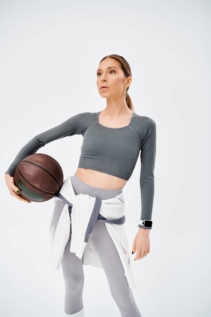 Спортивная молодая женщина в активной одежде уверенно держит баскетбол в правой руке на сером фоне. - Фото, изображение