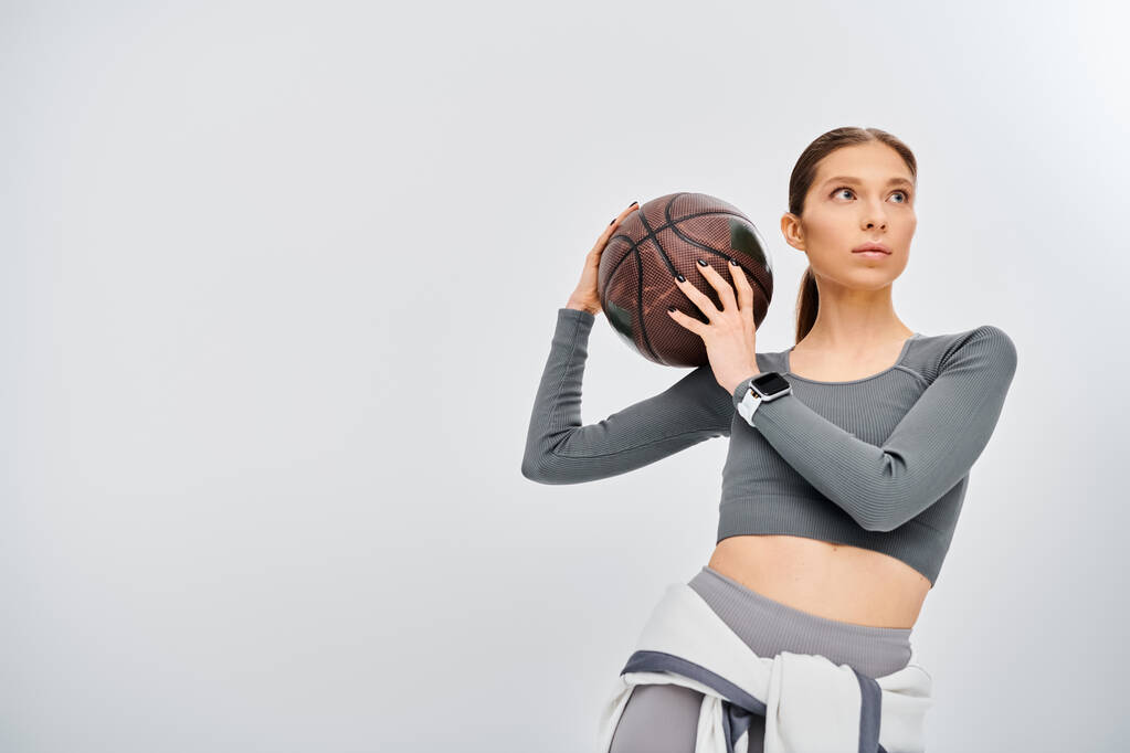 Спортивная молодая женщина держит баскетбольный мяч в правой руке, демонстрируя грацию и силу на сером фоне. - Фото, изображение