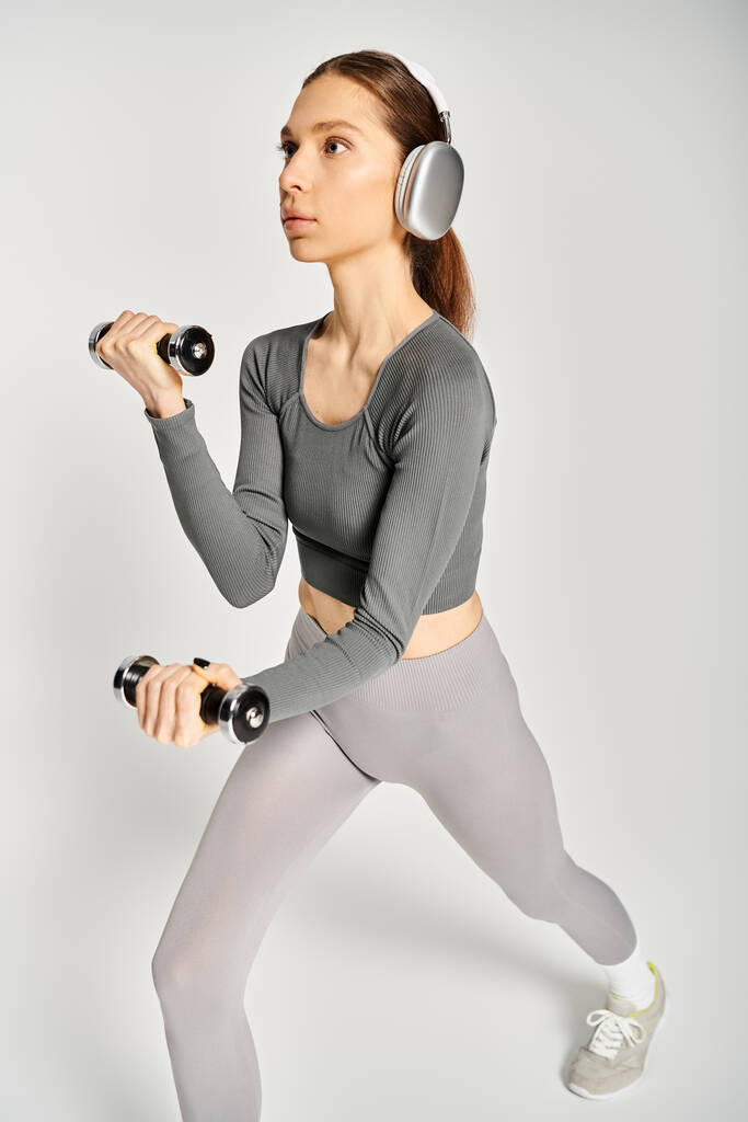 Egy sportos fiatal nő aktív kopás gyakorlatok súlyzókkal, miközben zenét hallgat fülhallgatón keresztül, szürke háttérrel. - Fotó, kép