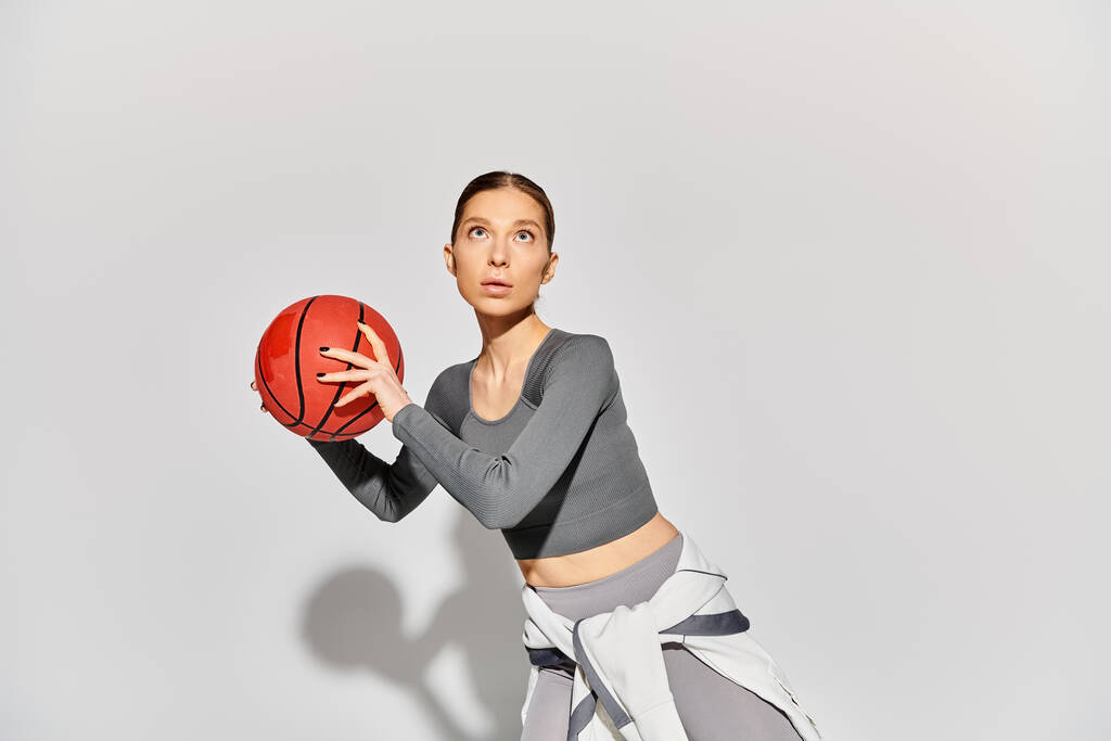 Una joven deportista en ropa activa sosteniendo una pelota de baloncesto en su mano derecha sobre un fondo gris. - Foto, imagen