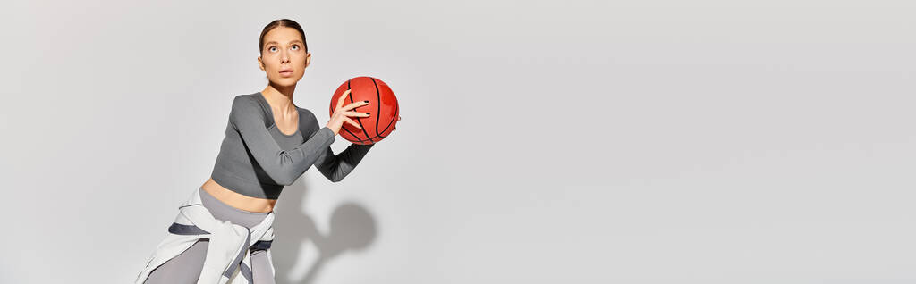 Une jeune femme sportive en tenue active tenant un ballon de basket rouge dans sa main droite sur un fond gris. - Photo, image