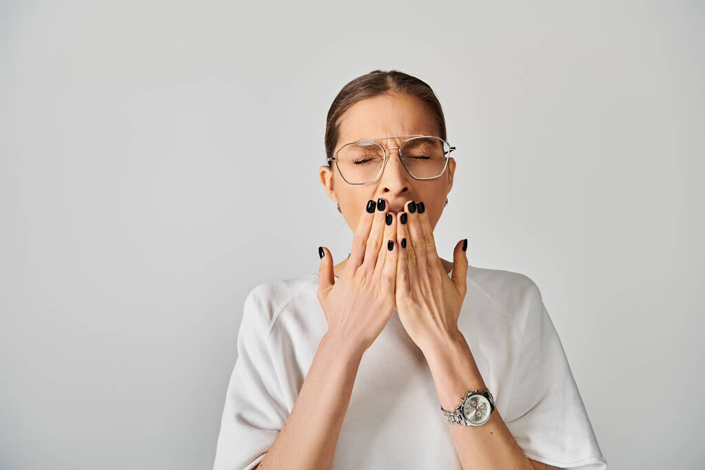 Eine junge Frau in weißem T-Shirt und Brille, die den Mund mit den Händen vor grauem Hintergrund verdeckt. - Foto, Bild