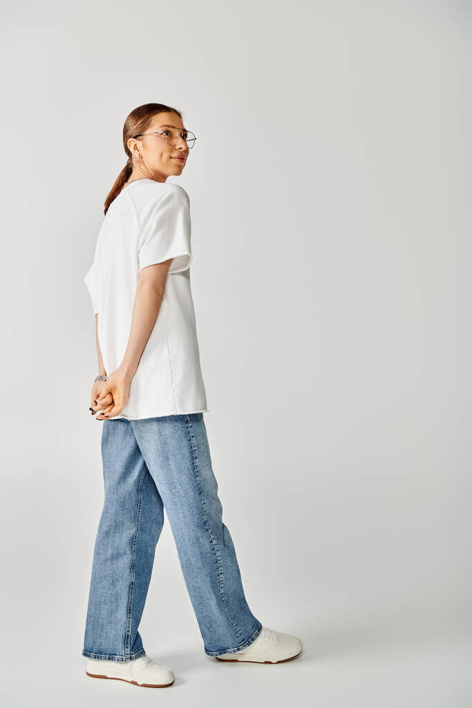 Uma jovem mulher de camisa branca e jeans caminha graciosamente sobre um fundo cinza. - Foto, Imagem