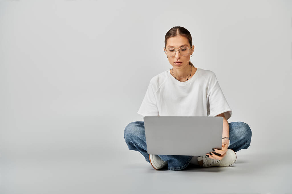 Una joven con una camiseta blanca y gafas sentadas en el suelo, absorta en su portátil sobre un fondo gris. - Foto, imagen
