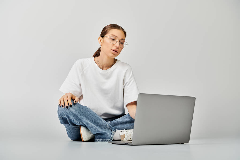 A young woman in a white t-shirt and glasses sits on the floor, focused on her laptop screen, engaged in work or study. - Photo, Image