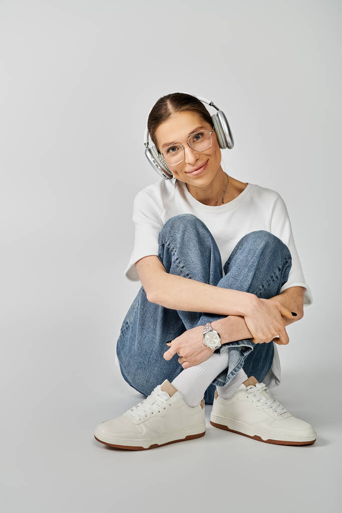Una joven con una camiseta blanca y gafas se sienta en el suelo, sumergida en su música mientras usa auriculares. - Foto, imagen