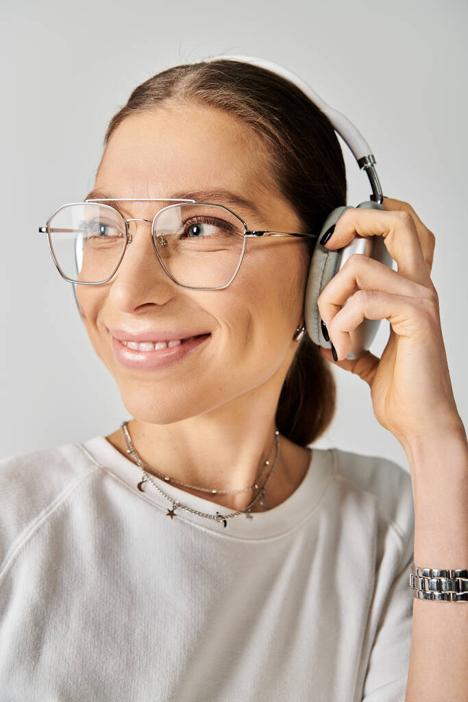 Egy fiatal nő fehér pólóban és szemüvegben fülhallgatót tart szürke alapon.. - Fotó, kép