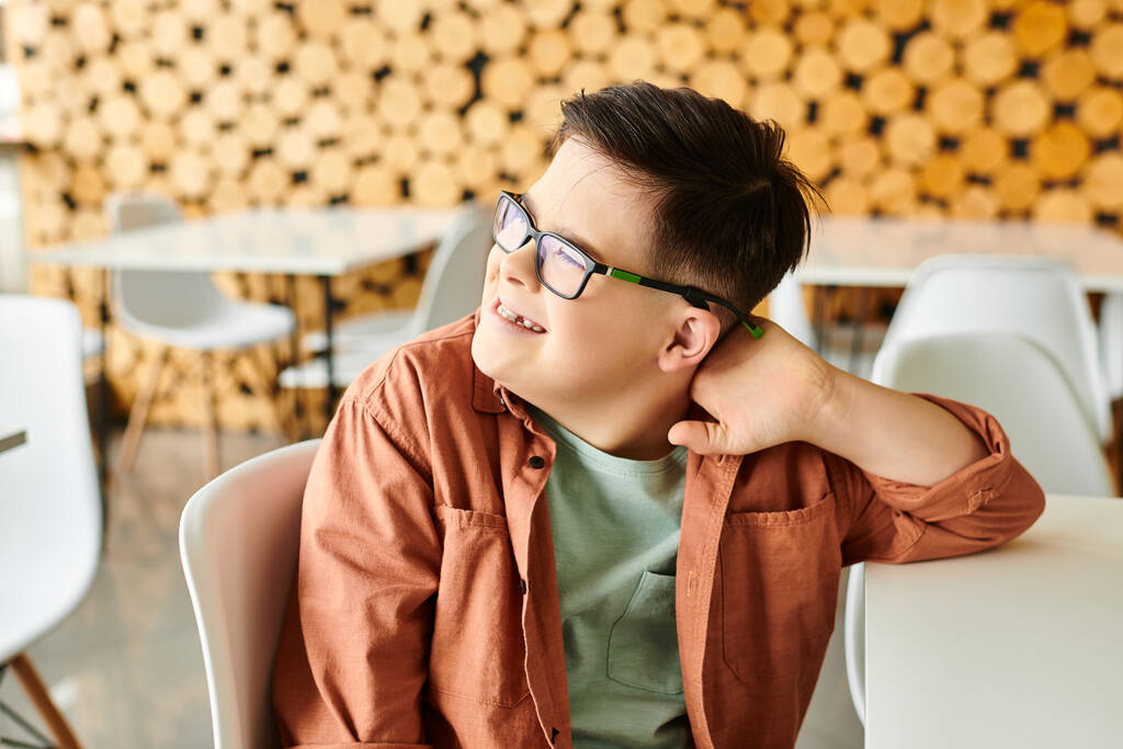 χαρούμενο προεφηβικό αγόρι χωρίς αποκλεισμούς με σύνδρομο Down σε casual ενδυμασία κοιτάζοντας μακριά ενώ στο καφέ - Φωτογραφία, εικόνα
