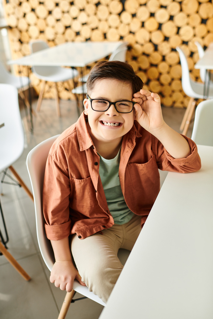 alegre inclusivo preadolescente chico con síndrome de Down con gafas sentado en la cafetería y sonriendo a la cámara - Foto, imagen