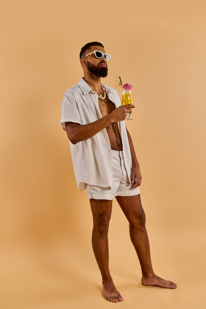 Ένας κομψός άντρας με λευκό πουκάμισο και σορτς απολαμβάνει ένα δροσιστικό ποτό σε ένα γαλήνιο περιβάλλον, αποπνέοντας χαλάρωση και ικανοποίηση. - Φωτογραφία, εικόνα