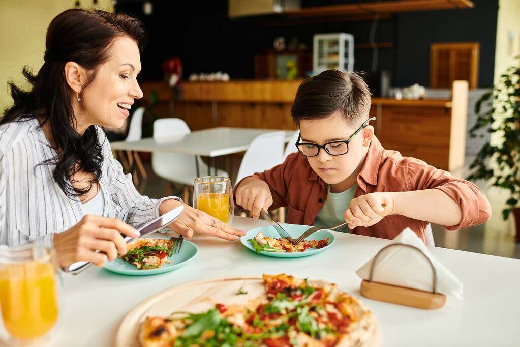 alegre madre comer pizza y beber jugo con su inclusivo lindo hijo con síndrome de Down - Foto, imagen