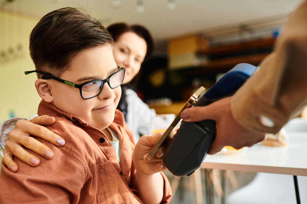 schattig inclusief jongen met Down syndroom betalen met smartphone in cafe in de buurt van zijn vrolijke moeder - Foto, afbeelding