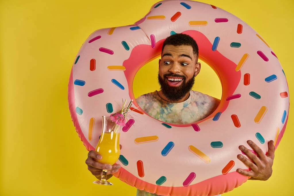 Ένας άντρας που κρατάει με χαρά ένα τεράστιο ντόνατ και ένα δροσιστικό ποτό, δείχνοντας αγάπη για νόστιμες λιχουδιές και χαλάρωση. - Φωτογραφία, εικόνα