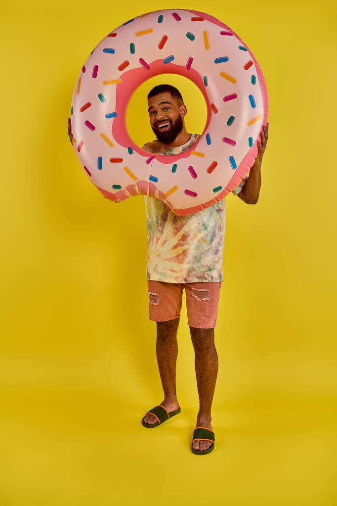 Ένας άντρας κρατά παιχνιδιάρικα ένα τεράστιο ντόνατ μπροστά στο πρόσωπό του, καλύπτοντας το εντελώς. Τα πολύχρωμα τρούφα έρχονται σε αντίθεση με την έκφραση της χαράς του. - Φωτογραφία, εικόνα