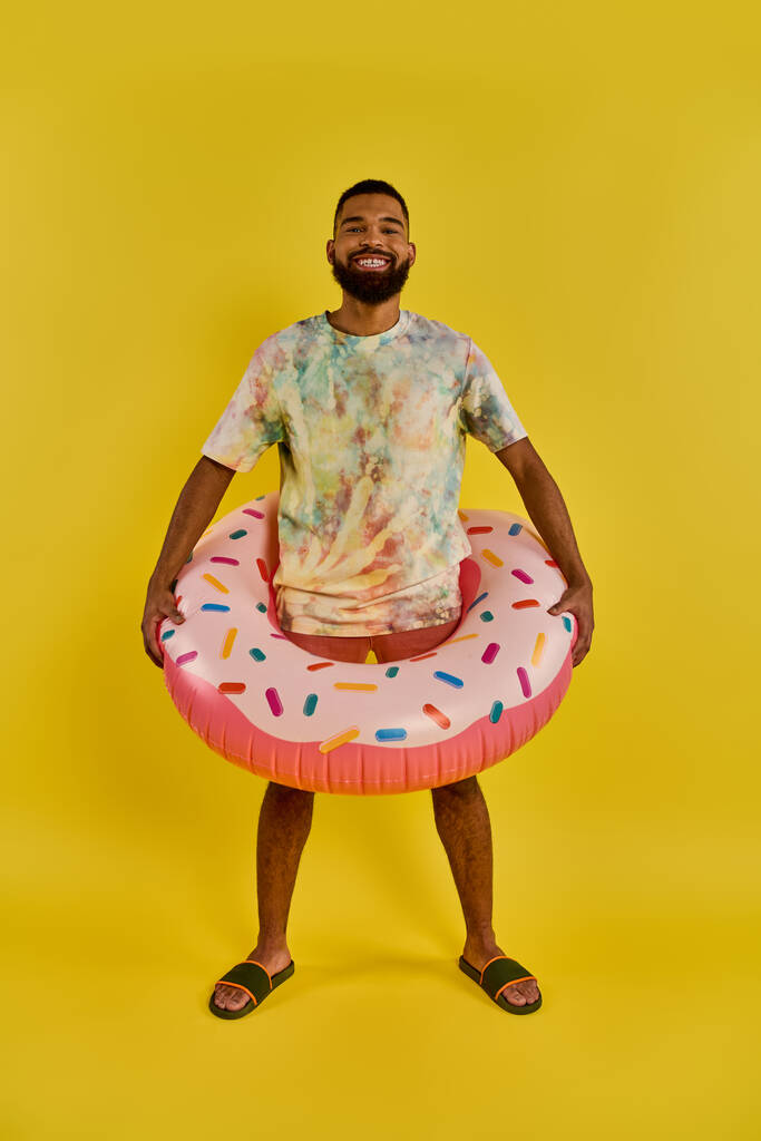 Un homme tient joyeusement un anneau gonflable en forme de donut coloré, contrastant avec un ciel bleu ensoleillé, incarnant détente et plaisir au bord de l'eau. - Photo, image