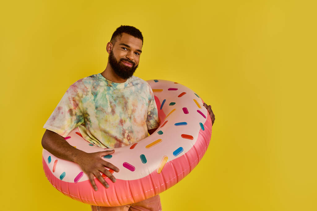 Ein Mann hält freudig einen riesigen Donut vor einem leuchtend gelben Hintergrund und präsentiert die zuckerhaltige Leckerei. - Foto, Bild