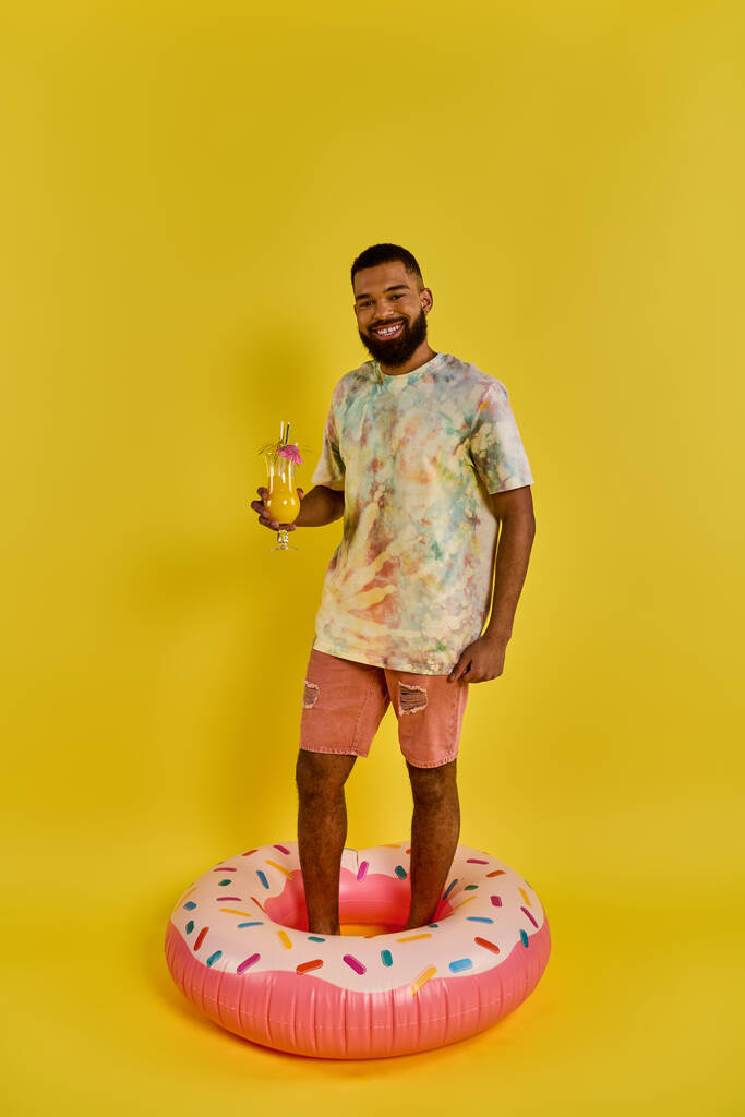 Ein Mann in legerer Kleidung steht auf einem bunten Donut-förmigen Schwimmer in einem Pool, hält ein Getränk in der Hand und genießt den Moment. - Foto, Bild