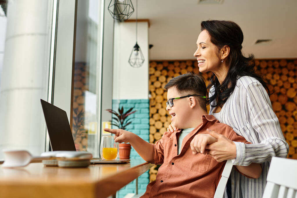 радостная мать проводит время со своим инклюзивным сыном с синдромом Дауна перед ноутбуком в кафе - Фото, изображение