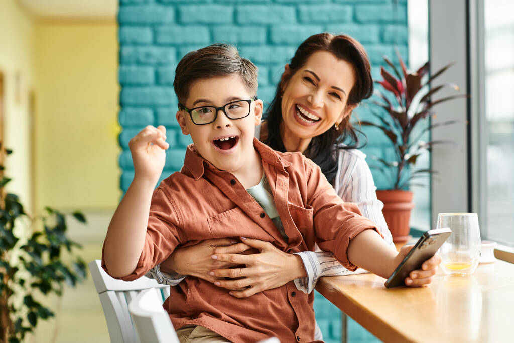 veselý inkluzivní preteen chlapec s Downovým syndromem s úsměvem na kameru, zatímco drží telefon v blízkosti své matky - Fotografie, Obrázek
