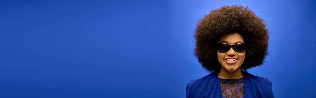 Trendige Afroamerikanerin mit lockigem Haar und Sonnenbrille, die eine blaue Jacke trägt und in Pose tritt. - Foto, Bild