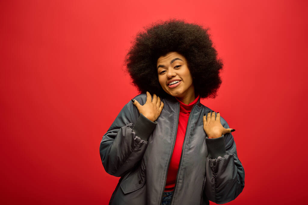 Stylish African American woman with curly hairdo posing in trendy attire against a vibrant red backdrop. - Photo, Image