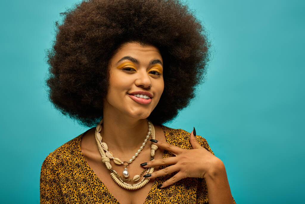 Стильна афроамериканська жінка з кучерявим волоссям у намисті, позує на яскравому тлі. - Фото, зображення