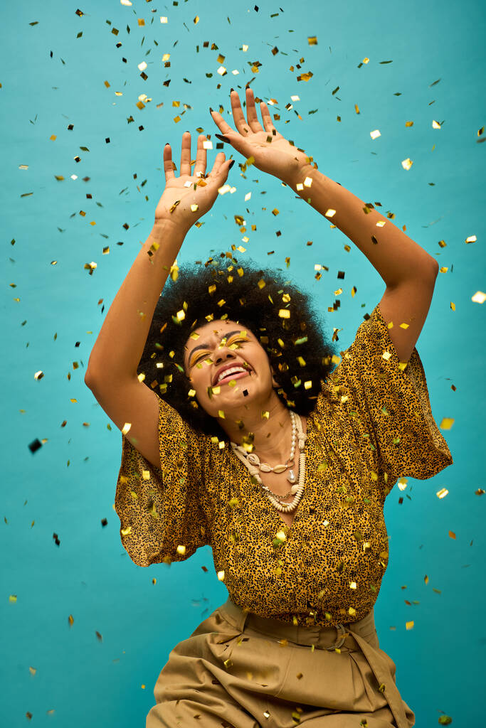 Nuori afroamerikkalainen nainen tyylikkäässä puvussa nostaa kädet, värikkäiden konfettien ympäröimänä.. - Valokuva, kuva