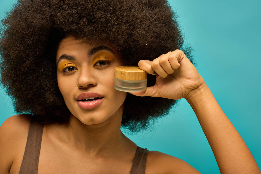 Стильна афроамериканська жінка з кучерявим зачіскою позує з баночкою макіяжу. - Фото, зображення