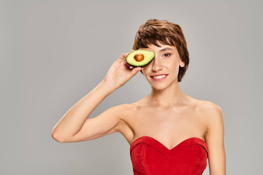 Jonge vrouw in een rode jurk poseert met een avocado tegen een levendige achtergrond. - Foto, afbeelding