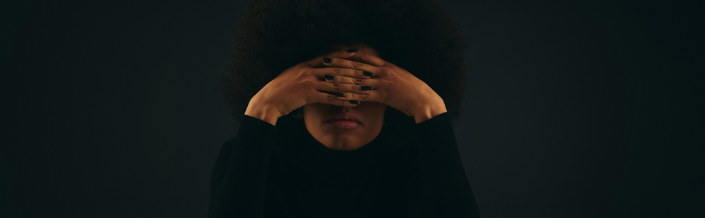Μια κομψή Αφροαμερικανή με μοντέρνα ενδυμασία καλύπτει το πρόσωπό της με τα χέρια της. - Φωτογραφία, εικόνα