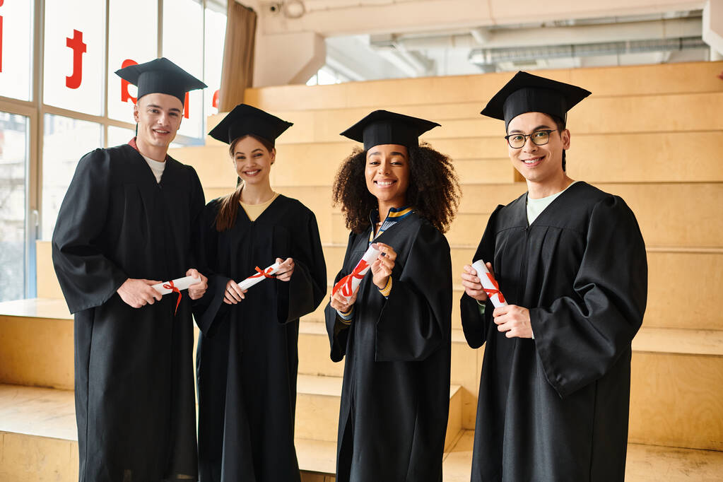 zróżnicowana grupa studentów w sukniach absolwentów pozujących z czapkami akademickimi i dyplomami - Zdjęcie, obraz