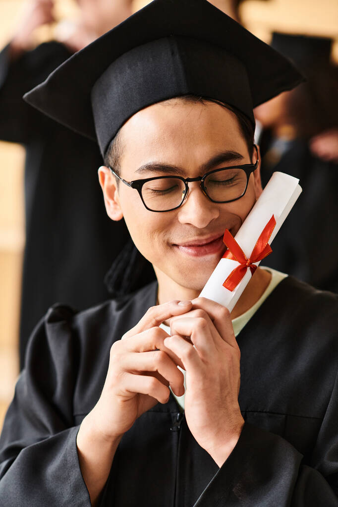 Asijský muž nosí maturitní čepici a šaty s úsměvem, zatímco drží diplom v ruce. - Fotografie, Obrázek