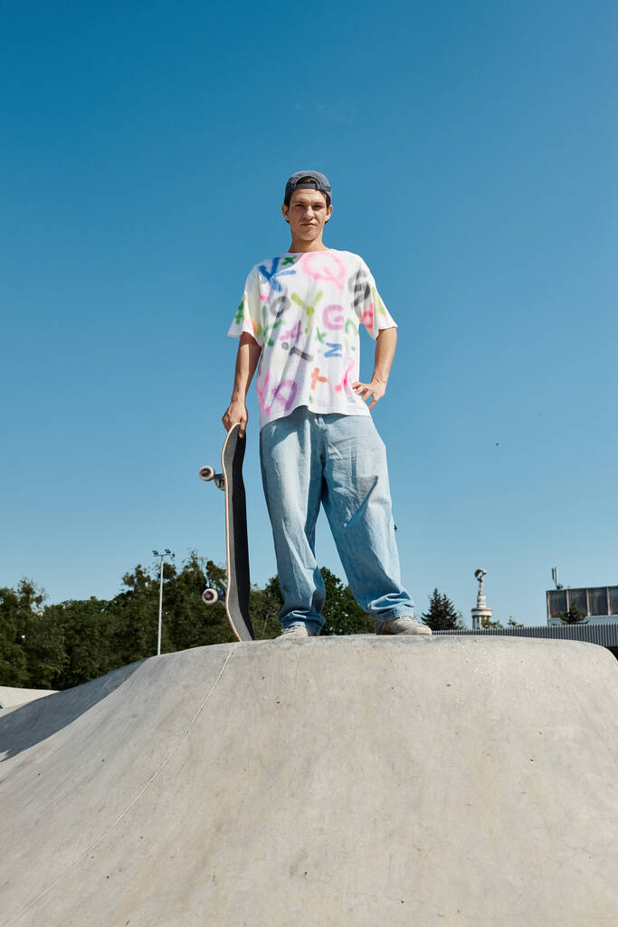 Ένας νεαρός σκέιτερ με αυτοπεποίθηση κατέχει ένα skateboard στην κορυφή μιας ράμπας σε ένα ζωντανό υπαίθριο πάρκο skate σε μια ηλιόλουστη μέρα του καλοκαιριού. - Φωτογραφία, εικόνα
