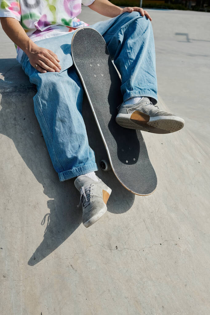 Ένα νεαρό αγόρι σκέιτερ κάθεται με σιγουριά σε ένα skateboard, έτοιμο να γλιστρήσει σε μια ράμπα σε ένα υπαίθριο πάρκο skate σε μια ηλιόλουστη μέρα του καλοκαιριού. - Φωτογραφία, εικόνα