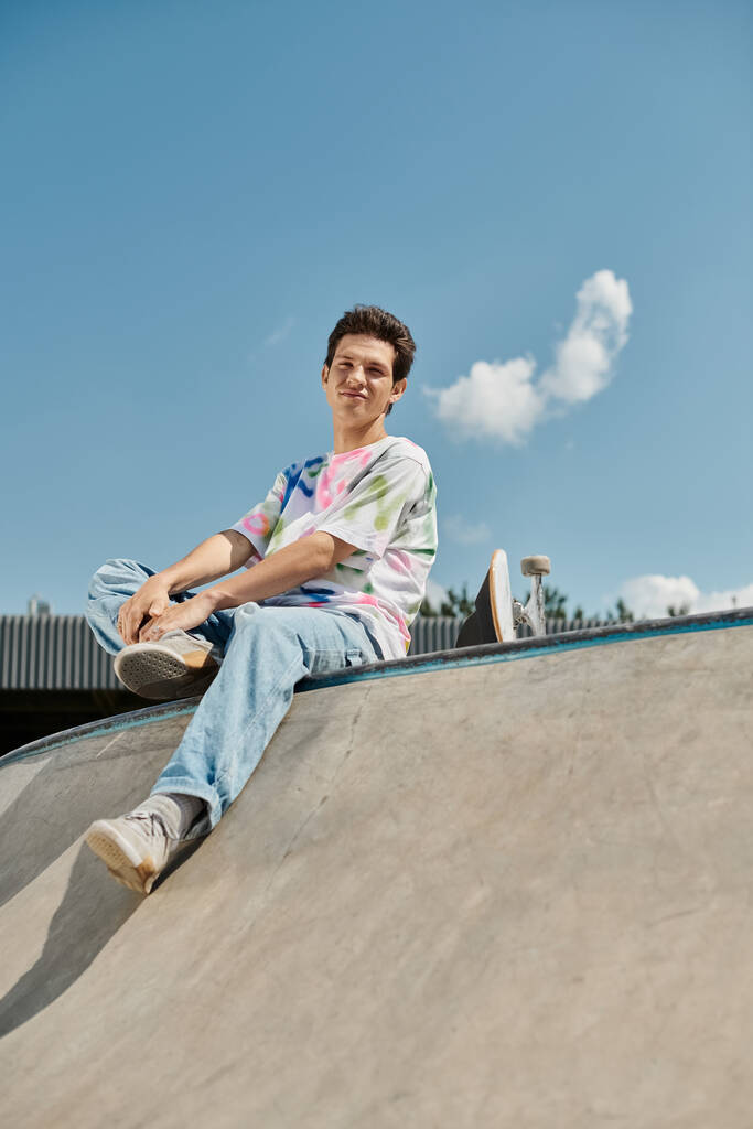 A young skater boy sits calmly at the top of a skateboard ramp in a vibrant outdoor skate park on a sunny summer day. - Photo, Image