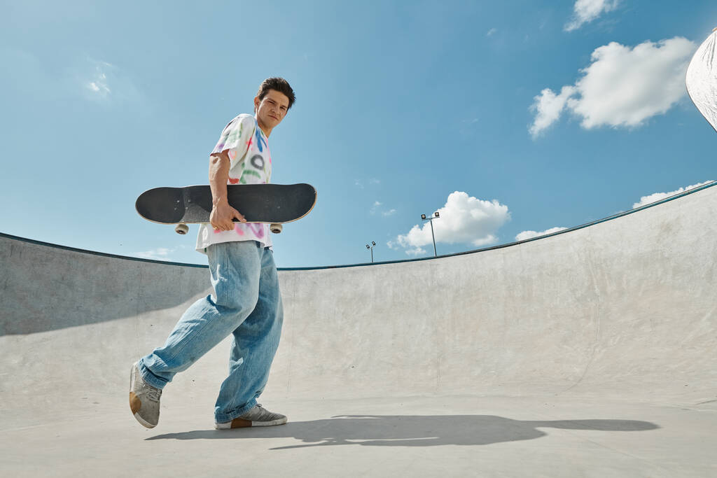 Un joven caminando con confianza en un parque de skate, sosteniendo su monopatín bajo el sol brillante. - Foto, Imagen