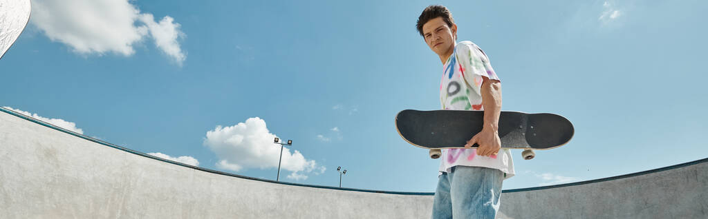 Un giovane uomo tiene uno skateboard, in piedi in un parco skate vibrante in una giornata di sole, trasudando un senso di stile e libertà. - Foto, immagini