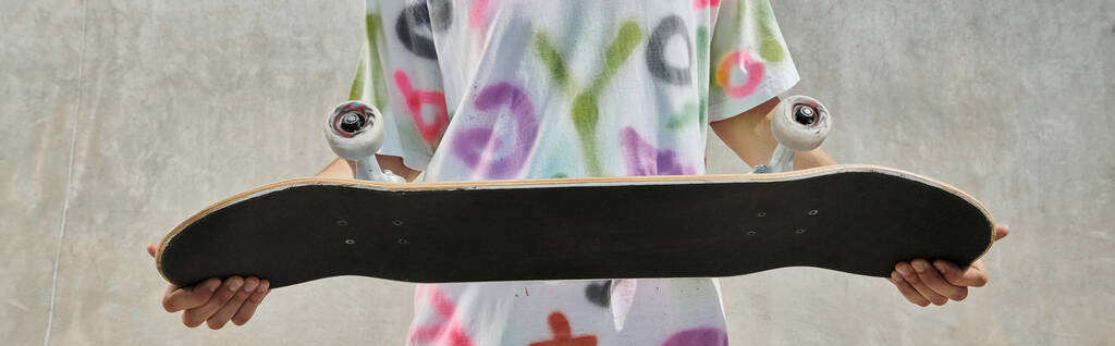 Ένα νεαρό αγόρι skateboard κρατώντας ένα skateboard σε ένα ηλιόλουστο πάρκο skate, προετοιμασία για μια συναρπαστική βόλτα σε μια καλοκαιρινή μέρα. - Φωτογραφία, εικόνα