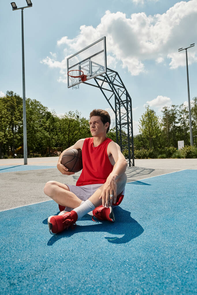 Ein junger Basketballspieler sitzt an einem sonnigen Sommertag gedankenverloren auf dem Platz und hält einen Basketball in der Hand. - Foto, Bild