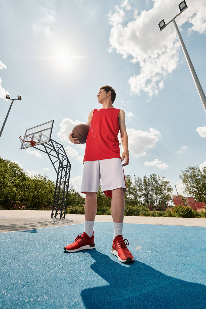 Ένας νεαρός μπασκετμπολίστας στέκεται στο γήπεδο, κρατώντας ένα μπάσκετ, έτοιμος να παίξει στην καλοκαιρινή ζέστη. - Φωτογραφία, εικόνα