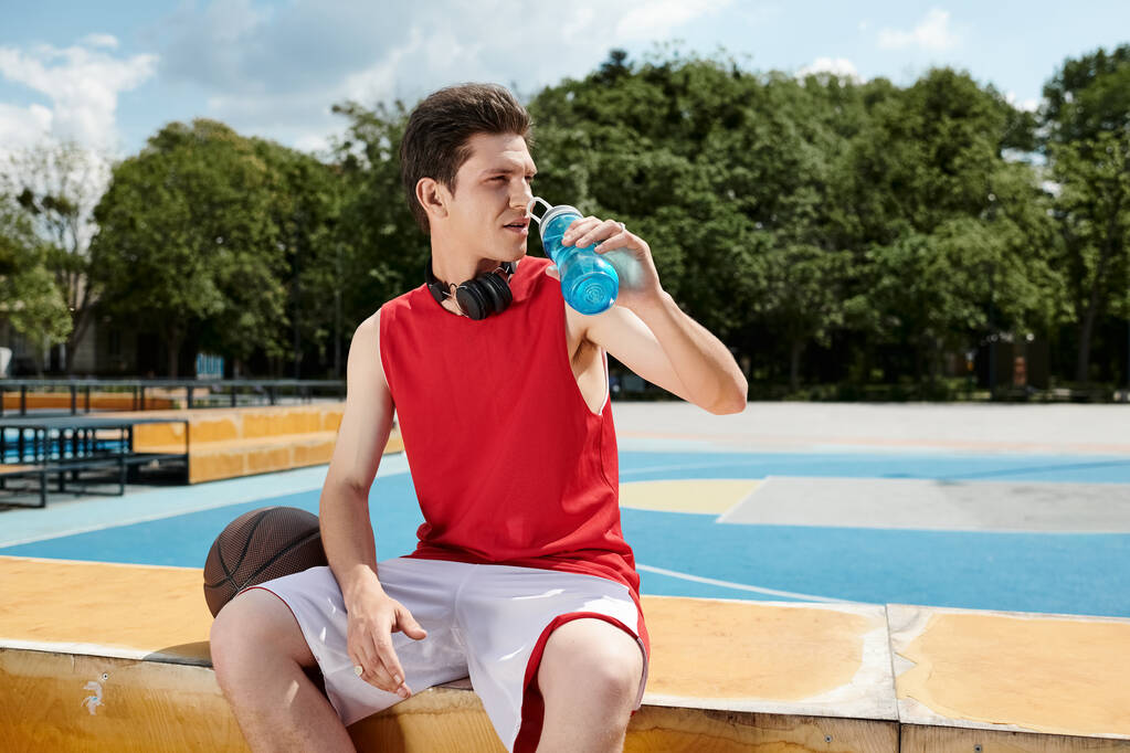 Ein junger Mann sitzt auf einem Sims und trinkt an einem sonnigen Tag aus einer Wasserflasche. - Foto, Bild