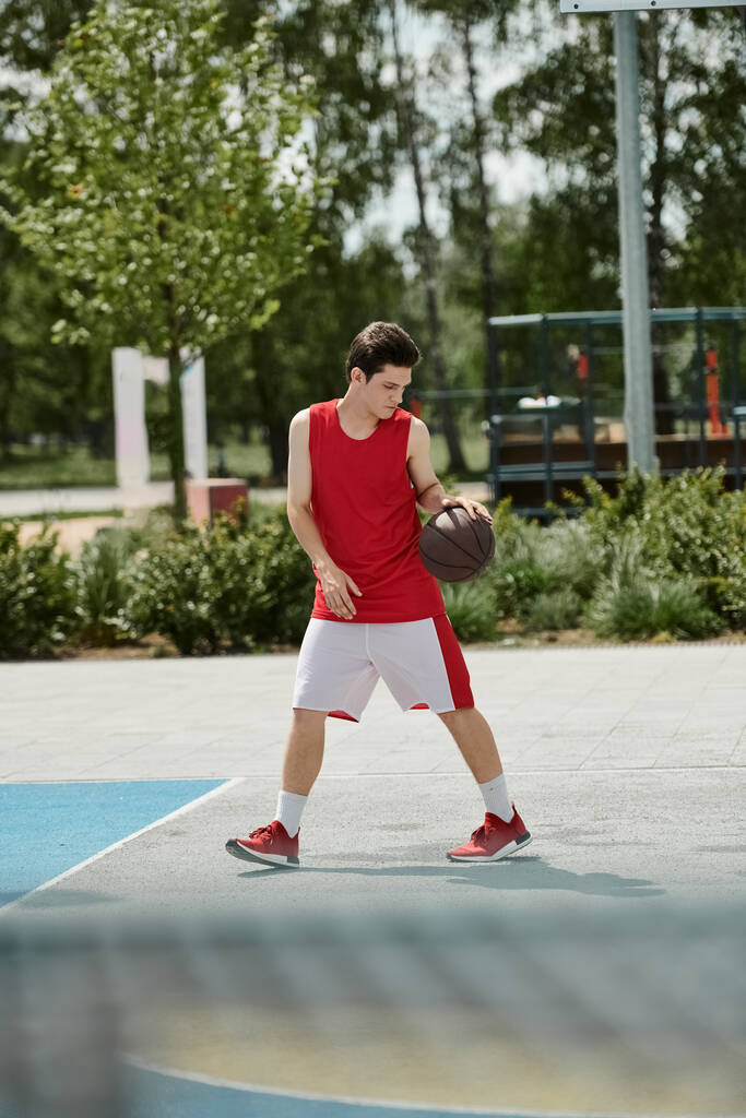Un jeune homme se tient sur un terrain de basket, tenant une balle dans sa main, prêt à jouer sous le soleil d'été. - Photo, image