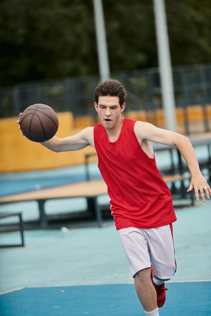 Ein junger Mann hält an einem sonnigen Tag selbstbewusst einen Basketball auf einem lebhaften Basketballfeld. - Foto, Bild