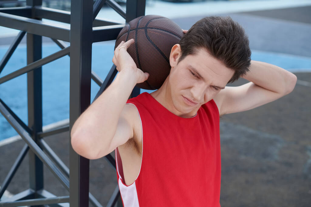 Młody mężczyzna w czerwonej koszuli umiejętnie trzyma koszykówkę podczas przygotowań do gry na świeżym powietrzu w słoneczny letni dzień. - Zdjęcie, obraz