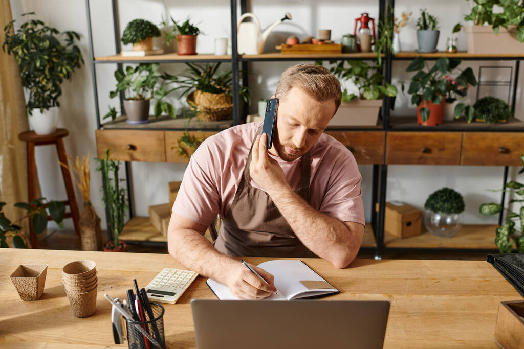 Un hombre se sienta en un escritorio hablando en un teléfono celular mientras trabaja en una tienda de plantas. - Foto, imagen