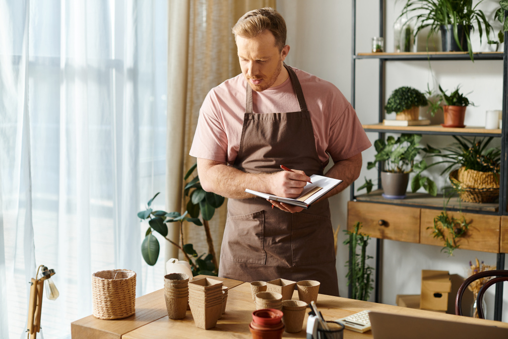 Un hombre en un delantal toma notas diligentemente en un portapapeles en una tienda de plantas, mostrando su dedicación a su pequeña empresa. - Foto, Imagen