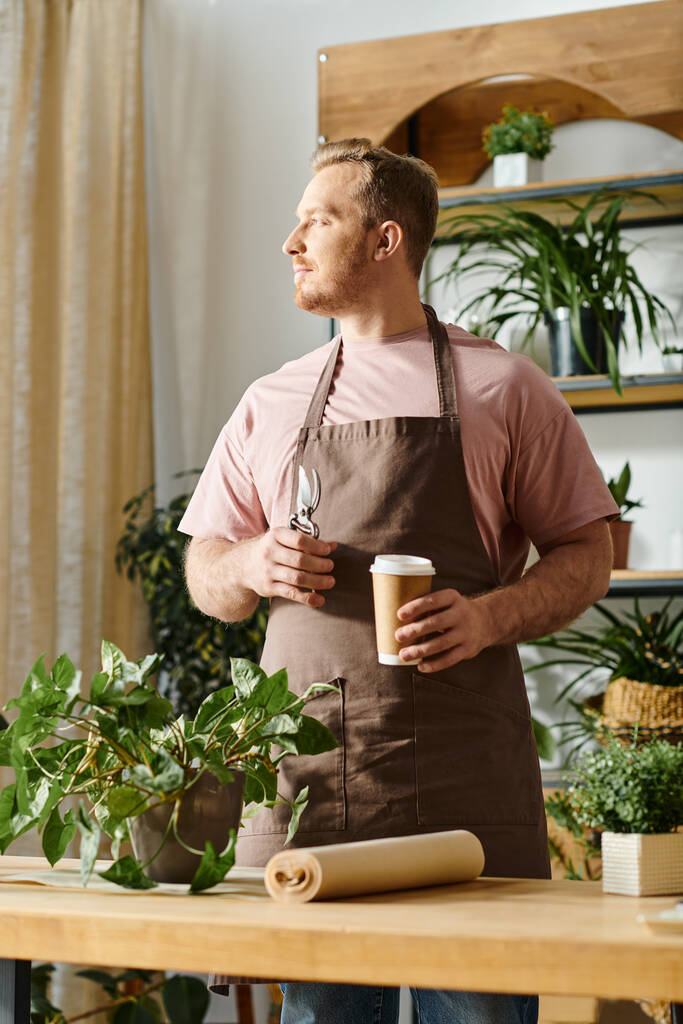 Ein Mann in Schürze genießt einen Moment der Entspannung und hält eine Tasse Kaffee in einem Pflanzenladen. - Foto, Bild