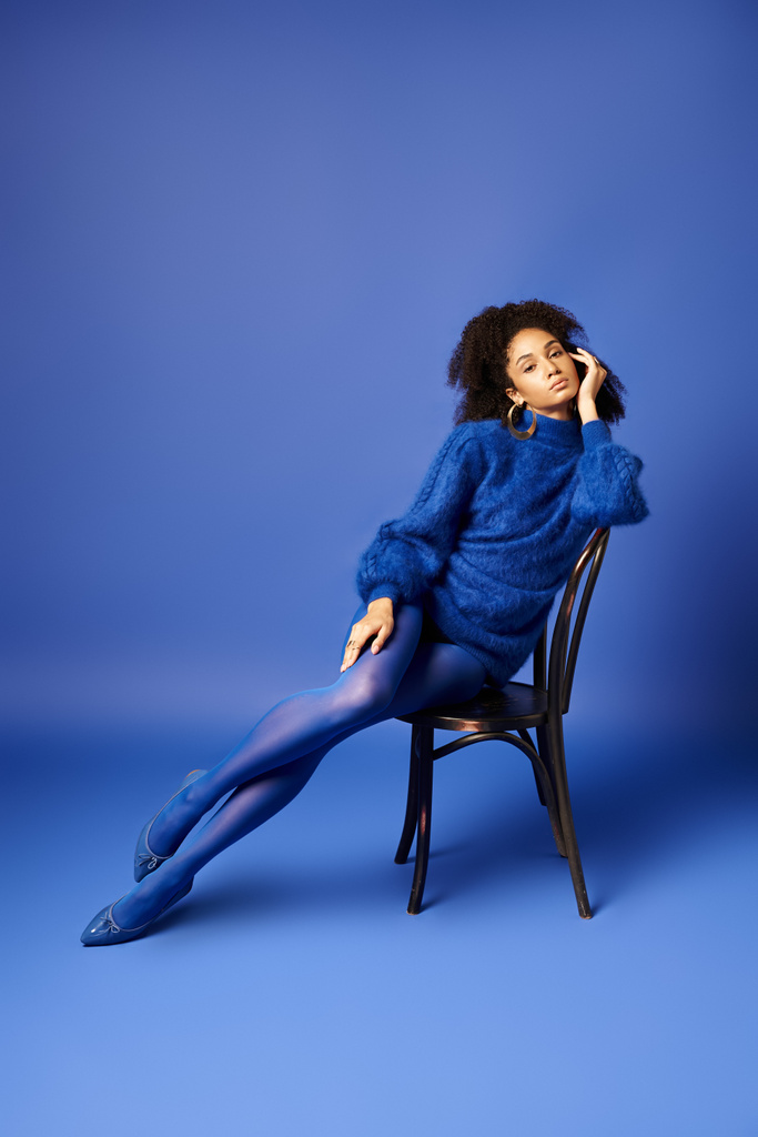 Молода жінка в стильній блакитній сукні елегантно сидить на стільці на яскравому синьому студійному фоні. - Фото, зображення