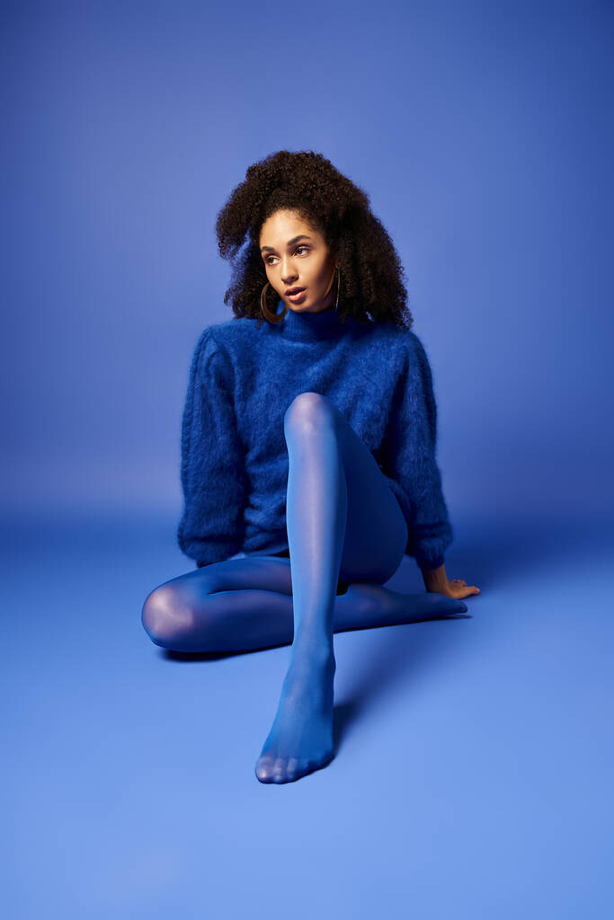 Una giovane donna elegante in calzamaglia vibrante e maglione siede a gambe incrociate su un pavimento blu studio, trasudando serenità e portamento. - Foto, immagini