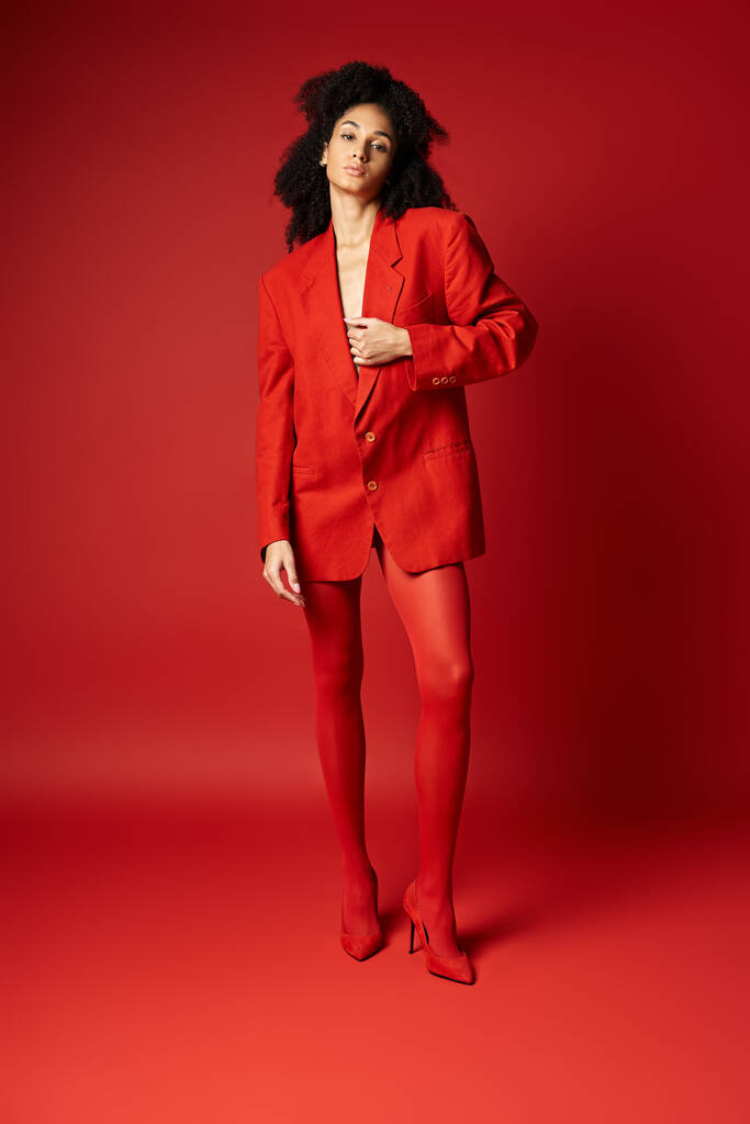 スタジオの設定でカメラのポーズを打つ鮮やかな赤いスーツのスタイリッシュな若い女性. - 写真・画像